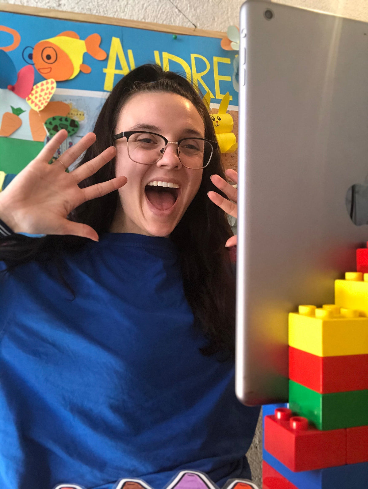 Smiling online English teacher holding a tablet beside Lego props ready for a young learner class